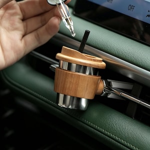 Op de afbeelding: Een auto-luchtverfrisser in de vorm van een miniatuur koffiebeker met een zilveren metalen behuizing, een lichtbruine houten huls en een bijpassend houten deksel. Een zwart staafje steekt uit de bovenkant en een glazen druppelaar hangt erboven.