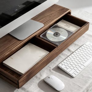 Puede incluir: Un soporte de madera de color marrón oscuro para monitor con un cajón de almacenamiento integrado. El cajón contiene una caja de CD de plástico transparente con un disco etiquetado Work Notes y un bloc de notas blanco sobre el soporte.
