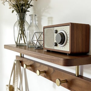 May include: A wooden shelf featuring a retro radio, glass vases, and a geometric metal ornament. Below, a row of gold-coloured hooks holds a tote bag. The shelf is mounted on a white wall.