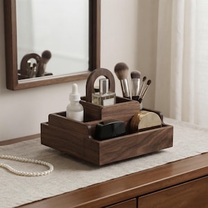 May include: A dark brown wooden organizer tray with a central handle, featuring multiple compartments holding a white dropper bottle, a clear glass perfume bottle, a black compact, a gold-toned container, and several makeup brushes.