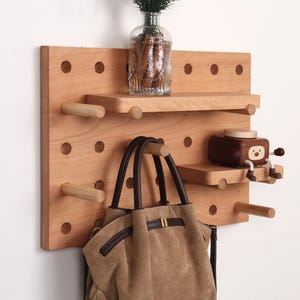 Op de afbeelding: Een lichtbruine houten pegboard wandorganizer met meerdere verwijderbare cilindrische pinnen, twee horizontale planken, een doorzichtige glazen vaas met groen en een kleine bruine, karaktervormige houder met witte ledematen.