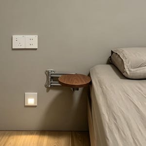 May include: A wall-mounted, circular wooden side table with a metal bracket. The table is attached to a grey wall next to a bed with a beige duvet and pillow. Electrical outlets and a light switch are also visible.