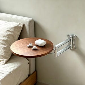 May include: A circular, dark wood bedside table mounted on a silver, adjustable arm. The table has a white charging case and two white earbuds. The table is attached to a wall next to a bed with a beige headboard.