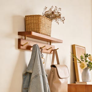 Op de afbeelding: Een houten wandplank met messing haken en een rieten mand. Een spijkerjas en een beige handtas hangen aan de haken. De plank is aan een crèmekleurige muur bevestigd, wat een stijlvolle opslag biedt.