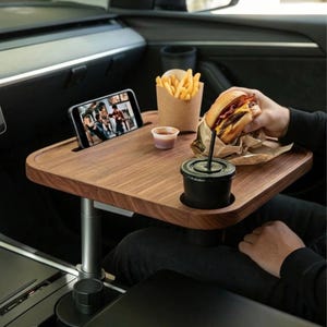 May include: A rectangular wooden car tray with a dark wood grain finish, featuring a built-in slot for a smartphone, a circular cup holder, and a smooth surface. The tray is mounted on an adjustable silver metal stand attached to the vehicle console.