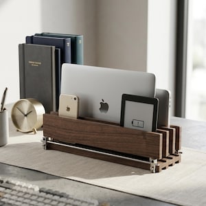 May include: A dark brown wooden vertical organiser stand featuring multiple slots for holding electronic devices, including a silver laptop, a gold smartphone, and a black e-reader with a low battery icon displayed on its screen.