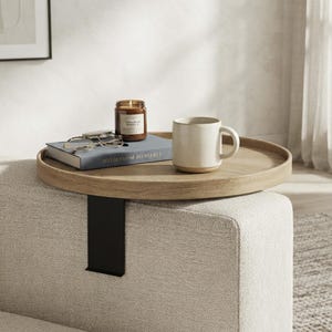 May include: A round light wood sofa arm tray table with a black metal clamp base. On the tray sits a blue hardcover book titled ESSAYS ON MINIMALISM, a pair of tortoiseshell glasses, a brown glass candle jar, and a cream ceramic mug.