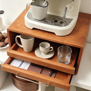 Può includere: Un organizer per caffè in legno con due cassetti estraibili e una superficie superiore che ospita una macchina per espresso color crema. Il cassetto superiore aperto contiene una tazza color crema, una tazzina su piattino e un bicchiere. Un libro intitolato Fredonia è all'interno.