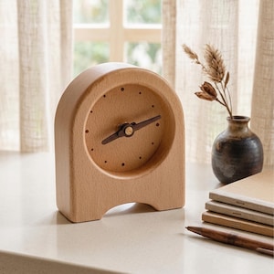 May include: A light-colored wooden table clock with an arched top and a curved base. The clock face features a smooth wood grain surface with small circular indentations for hour markers and two dark brown wooden hands at the center.