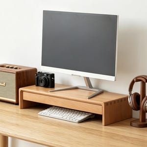 May include: A light-toned wooden monitor stand with a single drawer, featuring visible finger joint construction on the sides. The stand supports a silver-based computer monitor, with a white keyboard tucked underneath and a black camera on top.