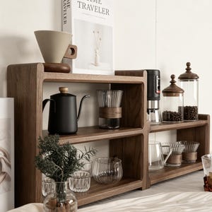 May include: A wooden coffee station with various coffee-making accessories. It includes a black gooseneck kettle, glass cups, a coffee grinder, and a pour-over coffee maker. The station also features glass jars with wooden lids and a decorative plant.