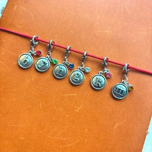 May include: Silver-toned charms with letters b, y, h, g, k, and m, each adorned with a small gemstone. The charms are suspended from a red cord, set against an orange background. These charms are ideal for jewelry making or crafting.