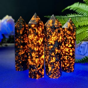 Puede incluir: Cuatro torres de cristal pulidas y verticales con un patr&oacute;n moteado de naranja y negro. Los cristales tienen una parte superior puntiaguda y est&aacute;n dispuestos sobre una superficie azul, con un fondo oscuro y algo de vegetaci&oacute;n.