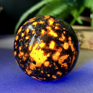 Puede incluir: Una esfera pulida con un llamativo patrón de inclusiones negras y naranja-amarillo vibrante. La gema parece brillar, creando un efecto visual cautivador. La esfera es un objeto decorativo, posiblemente para exhibición o colección.