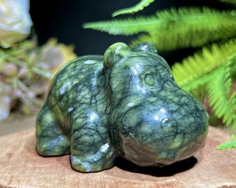 May include: A green stone hippo figurine with white veining. The hippo is carved in a rounded, stylized form, with visible eyes and a large snout. The figurine is placed on a wooden surface, with green foliage in the background.