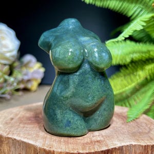 May include: A green stone sculpture of a female torso. The polished surface reflects light, highlighting the curves of the form. The sculpture is displayed on a wooden base, with greenery in the background.