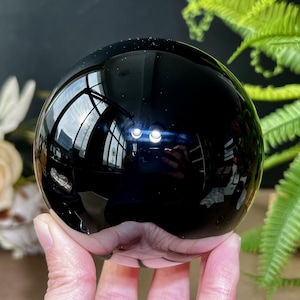 May include: A highly polished, black obsidian sphere. The surface reflects the surrounding environment, including a window and greenery. The sphere is held in a hand, with the smooth, glossy surface catching the light.