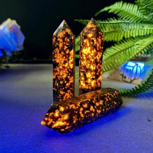 Puede incluir: Tres torres de piedras preciosas naranjas y negras con puntas puntiagudas. Las piedras preciosas tienen un patr&oacute;n moteado y una apariencia brillante e iluminada. Los cristales est&aacute;n dispuestos sobre una superficie azul con follaje verde en el fondo.