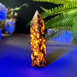 Puede incluir: Obelisco de cristal rectangular con punta. El cristal tiene una base oscura con inclusiones de color naranja y amarillo brillante. Se encuentra sobre una superficie azul, con follaje verde en el fondo. Aproximadamente 10 cm de altura.
