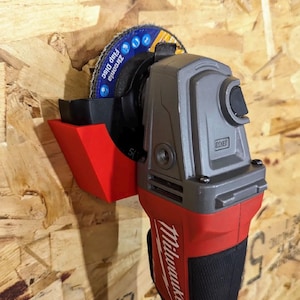 May include: A red and gray Milwaukee angle grinder with a blue and black flap disc attached. The grinder is mounted on a wooden surface. The brand name "Milwaukee" is visible on the tool.