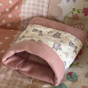 May include: A soft, cylindrical pet sleeping pouch made of solid dusty pink fleece fabric on the interior and edges, featuring a central band of cream-coloured fabric decorated with a repeating pattern of small grey rabbits and tiny floral accents.