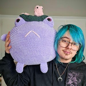 May include: A large, round, lavender crochet frog plushie with pink eyes, a green head covering, and a small pink creature on top. The plushie has a smiling face and small arms and legs.