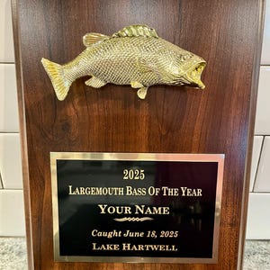 May include: A wooden plaque with a gold-colored largemouth bass sculpture mounted on top. The plaque has a black plate with gold lettering that reads "2025 Largemouth Bass Of The Year" and includes space for a name and the date and location of the catch.