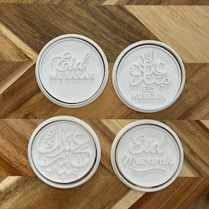 May include: Four white circular cookie cutters with the words "Eid Mubarak" in various fonts. The cutters are on a wooden surface with a diagonal grain pattern. The cutters are round with a raised border.