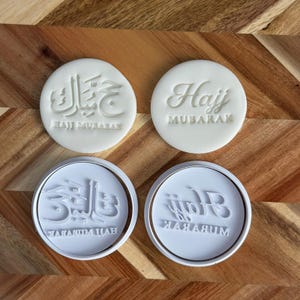 May include: Four white cookie stamps with text. Two stamps are round with the words "Hajj Mubarak" in a script font. The other two stamps have Arabic script and the words "Hajj Mubarak" in a smaller font.