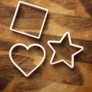 May include: Three white plastic cookie cutters: a square, a heart, and a star. They are arranged on a wooden surface with a brown, textured grain. Ideal for baking.
