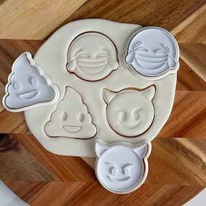 Pode incluir: Um conjunto de cortadores de biscoitos de plástico branco com cinco formas de emoji distintas, incluindo dois ícones de cocô, dois rostos a rir com lágrimas e um rosto de diabo, pressionados numa massa clara sobre uma superfície de madeira.