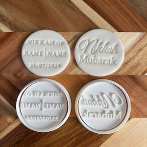 Peut inclure: Deux tampons à biscuits circulaires blancs et leurs impressions en relief correspondantes. Un tampon porte le texte NIKKAH OF NAME | NAME et 25/07/2026. L'autre tampon affiche le texte en écriture cursive Nikkah Mubarak. Fabriqués en plastique blanc.