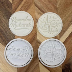 Puede incluir: Cuatro sellos redondos blancos para galletas con texto en relieve. Dos sellos dicen "Ramadan Mubarak" en inglés, y los otros dos presentan escritura árabe. Los sellos están sobre una superficie de madera.