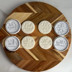May include: A selection of white cookie stamps featuring Arabic calligraphy and English phrases like "Eid Mubarak" and "Hajj Mubarak." They are displayed on a wooden board with a chevron design.