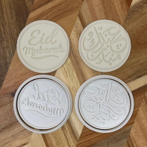 May include: Four white circular cookie cutters with embossed designs. Two cutters feature the words "Eid Mubarak" in English and the other two feature Arabic calligraphy. The cutters are on a wooden surface.