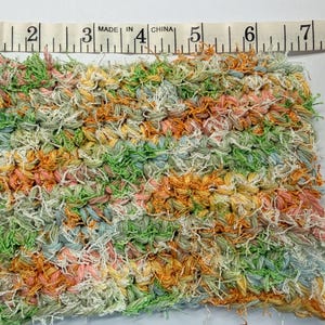 May include: A colorful, hand-woven textile square, approximately 6 inches wide, featuring a textured surface. The piece is made of yarn in shades of green, orange, pink, and blue. A measuring tape is placed above the textile.