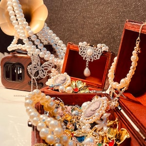 May include: A display of antique jewellery, including pearl necklaces, brooches, and earrings, presented in a wooden treasure chest. A decorative butterfly and a vintage-style radio are also visible. The jewellery features gold and silver tones with various gemstones.