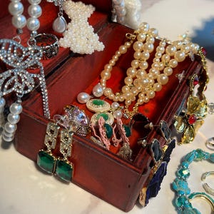 May include: A red wooden jewelry box overflowing with various jewelry pieces. The box contains pearl necklaces, earrings with emerald-green stones, and a beaded dress pendant. Other jewelry includes a silver ring and a turquoise bracelet.