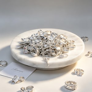 May include: A collection of silver rings, necklaces, and other jewelry pieces displayed on a white marble tray. The jewelry includes various ring designs, a key pendant necklace, and a butterfly ring. The brand name "Rinnar" is visible on a card.