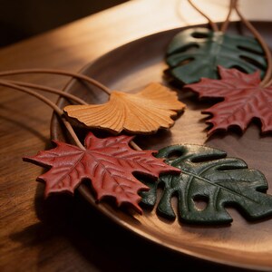 Peut inclure: Un ensemble d'ornements en cuir en forme de feuilles et de plantes, déclinés en rouge, vert et jaune. Chaque pièce présente des textures détaillées et est fixée à un fin cordon en cuir marron clair, formant des décorations aux formes naturelles.