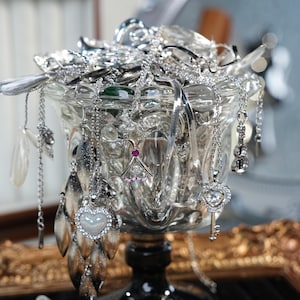 Könnte beinhalten: Eine Sammlung von silbernen Halsketten und Ohrringen, die in einer klaren, verzierten Glasschale ausgestellt sind. Der Schmuck umfasst Herz-, Schlüssel- und geometrische Anhänger. Die Schale steht auf einem schwarzen Sockel, mit unscharfem Hintergrund.