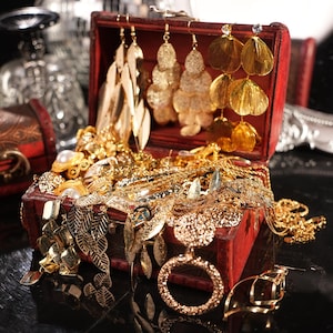 May include: A red wooden treasure chest overflowing with gold jewelry. The chest is open, revealing various necklaces, earrings, and rings. The earrings include leaf-shaped, tiered, and fan-shaped designs. The jewelry is displayed on a reflective surface.