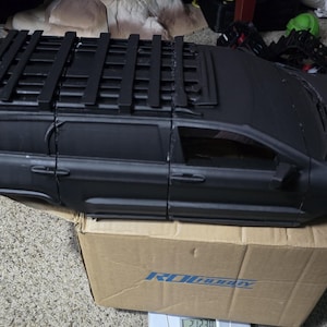 May include: A black model car body with a roof rack, sitting on a cardboard box. The car has detailed windows and doors. A red toy car is visible in the foreground.