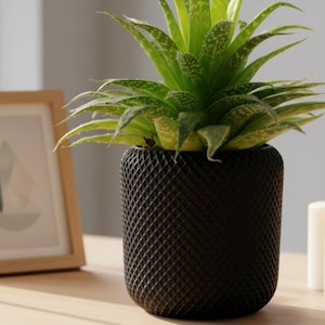 May include: A potted aloe vera plant with vibrant green leaves and white spots. The plant is in a black, textured pot with a diamond pattern. The pot sits on a wooden surface, with a framed picture and a white candle in the background.