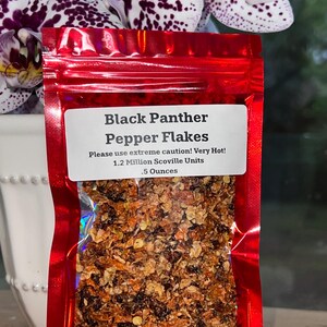May include: A red, shiny bag of Black Panther Pepper Flakes. The label reads "Please use extreme caution! Very Hot!" and lists 1.2 Million Scoville Units and .5 Ounces. The bag is filled with a mix of red and brown flakes. Purple and white orchids are in the background.