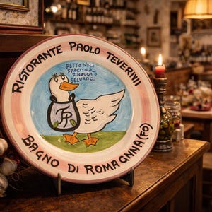 May include: A decorative ceramic plate with a duck illustration, featuring the text "Ristorante Paolo Teverini, Bagno di Romagna (Fo)". The plate is displayed on a wooden surface, with a restaurant interior in the background.