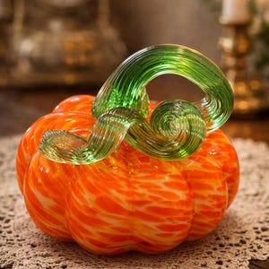 May include: A decorative glass pumpkin with an orange body and a green, swirled stem. The pumpkin has an orange and white speckled pattern. The glass pumpkin is a fall-themed decorative item.