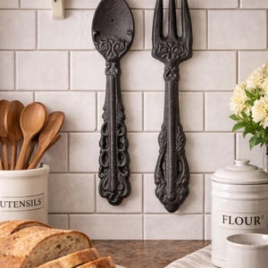 May include: Black cast iron spoon and fork wall decor with ornate detailing. The kitchen utensils are mounted on a white tiled wall. A loaf of sliced bread sits on a wooden cutting board, with a flour container and utensils nearby.
