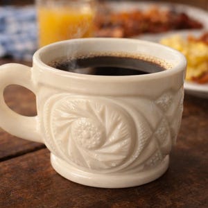 May include: A white ceramic coffee mug filled with dark coffee, with a decorative embossed pattern. The mug sits on a wooden surface, with breakfast items in the background, including bacon and scrambled eggs.