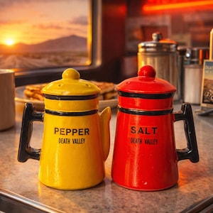 May include: A yellow "Pepper" and red "Salt" ceramic shakers with black handles and lids, featuring "Death Valley" text. The shakers sit on a diner counter with a coffee mug and a scenic window view.